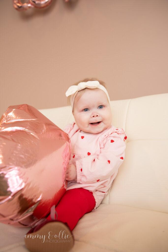 baby girl sits on white couch smiling at camera and playing with pink heart balloon