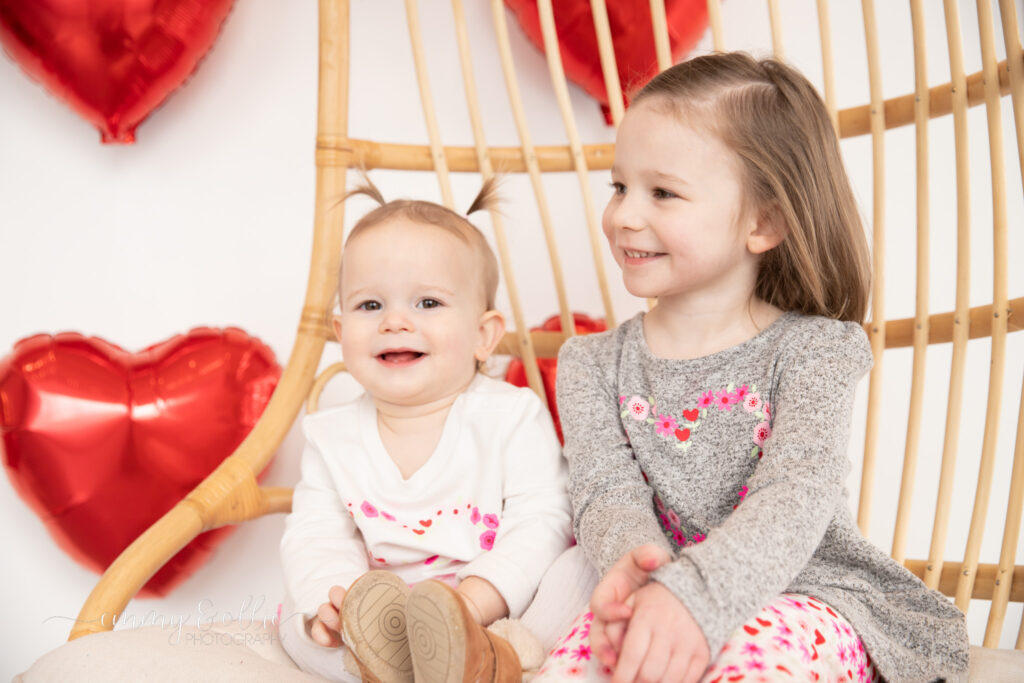 young sisters sit on decorative chair in front of wall decorated with red heart balloons for valentine's day and smile