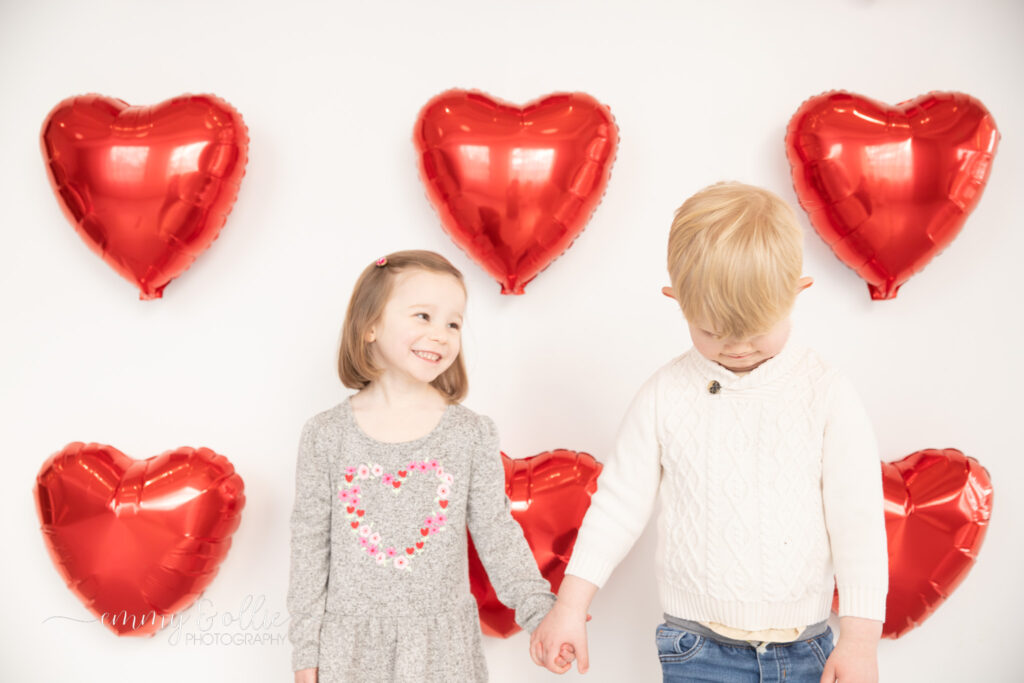 toddler girl and boy hold hands in front of white wall decorated with red heart balloons for valentine's day