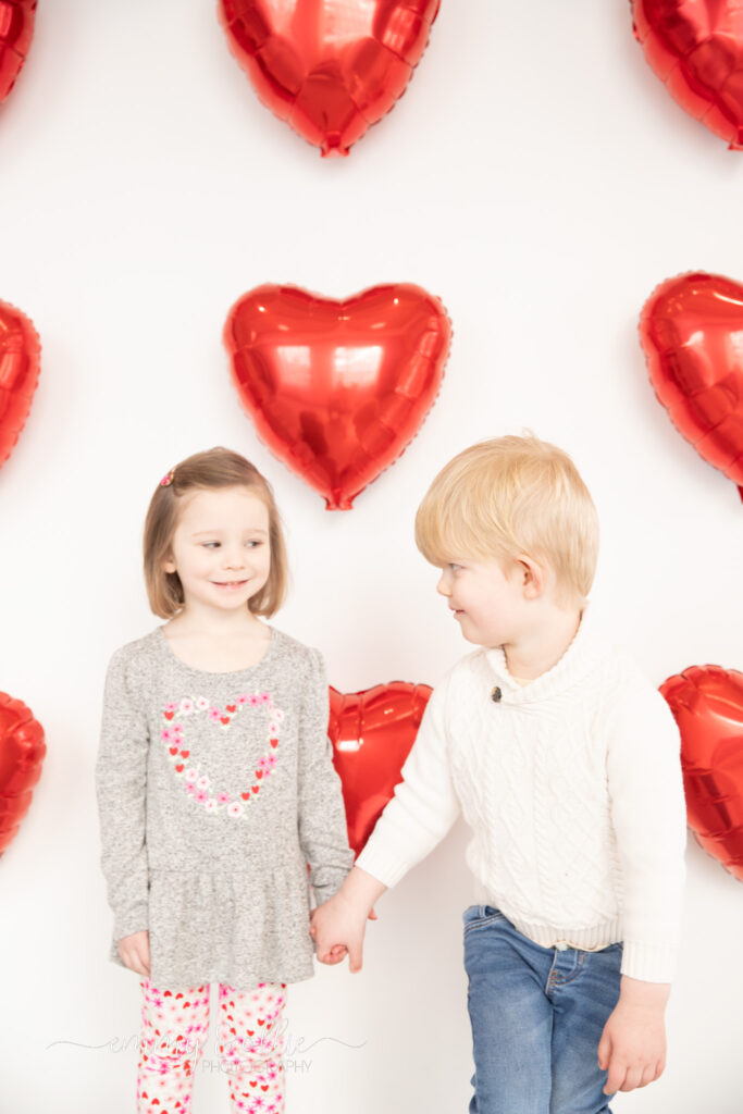 toddler girl and boy hold hands in front of white wall decorated with red heart balloons for valentine's day