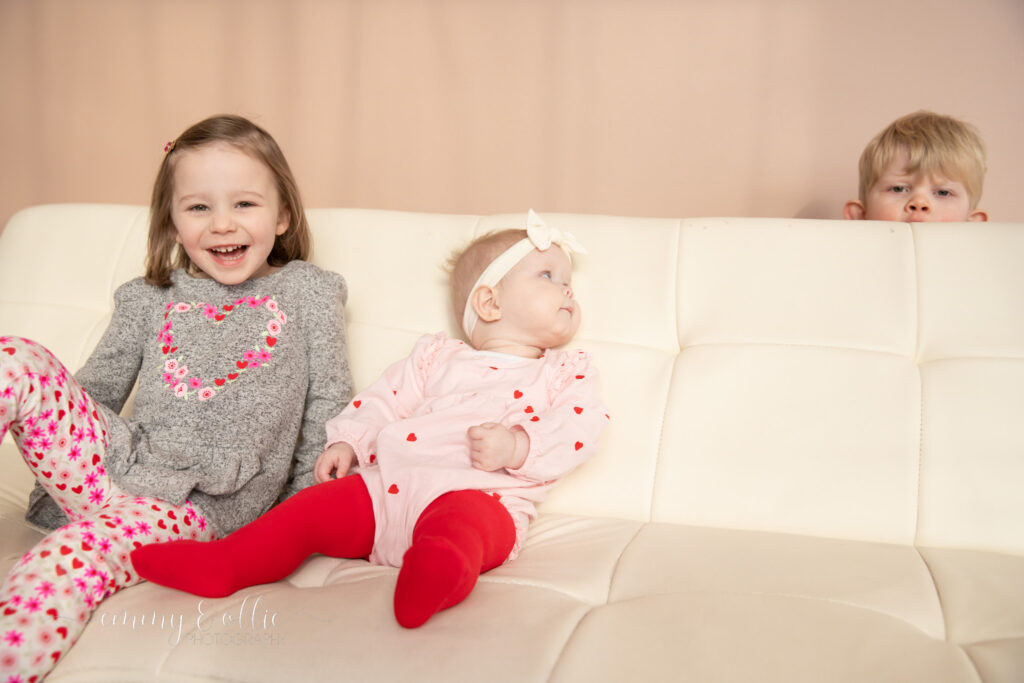 two young girls sit on white couch while toddler boy pops up behind couch