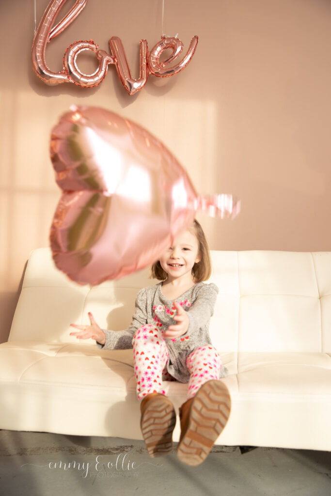 toddler girl smiles as pink heart balloon floats down in front of her as she sits on a white couch in front of a pink wall decorated with pink love balloon