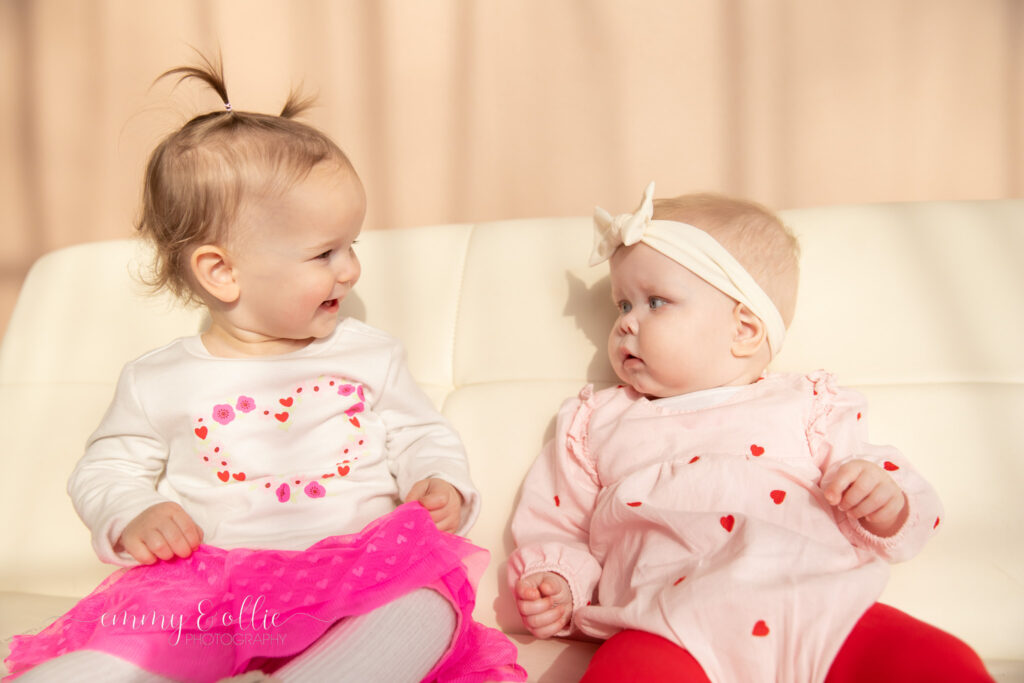 two baby girls sit and smile at each other on white couch in front of pink wall