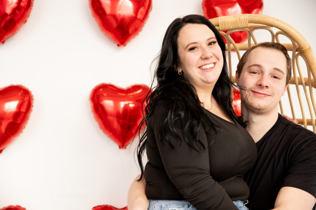 woman sits on boyfriend's lap in decorative chair in front of white wall decorated with red heart balloons