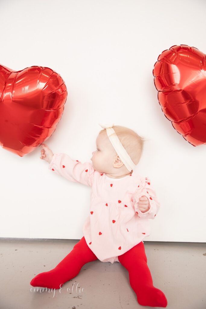 baby girl sits in front of white wall decorated with red heart balloons for valentine's day and reaches out for balloons