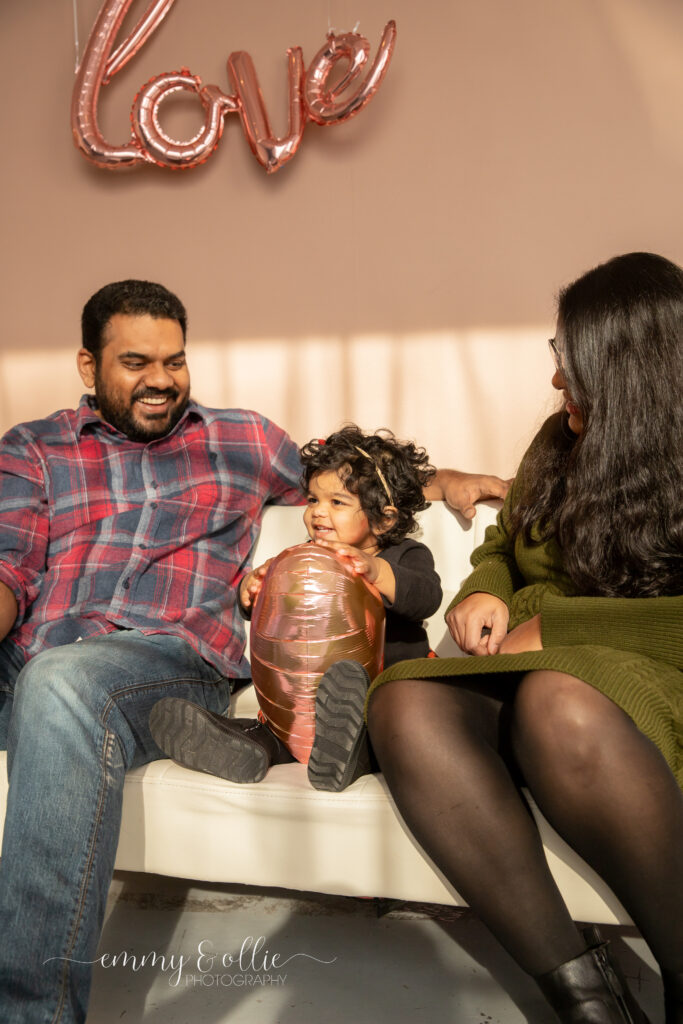 toddler girl holds pink heart balloon as parents look at her and smile