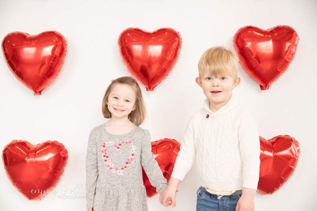 toddler girl and boy hold hands in front of white wall decorated with red heart balloons for valentine's day