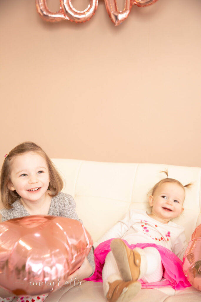 young sisters sit and laugh on white couch in front of pink wall while holding pink heart balloons