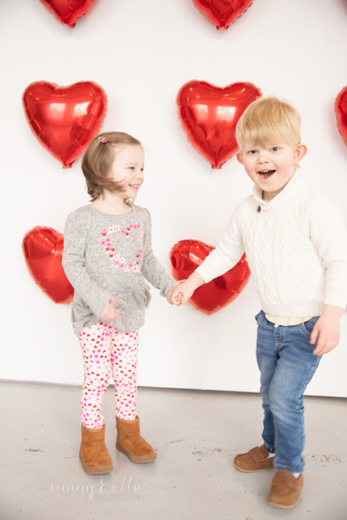 toddler girl and boy hold hands and dance in front of white wall decorated with red heart balloons for valentine's day