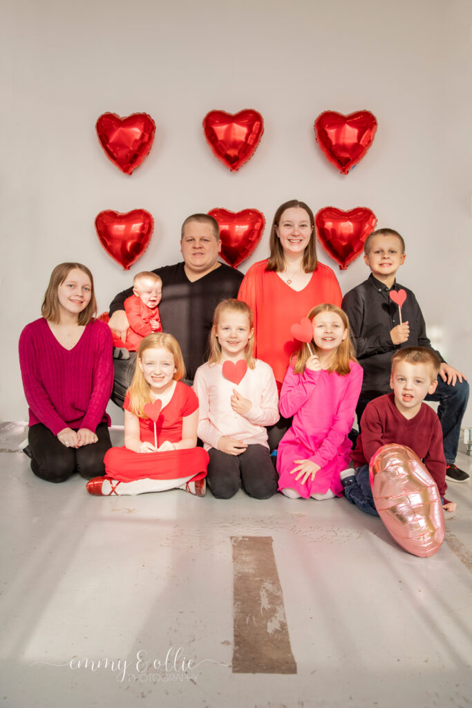 family sits on floor and poses in front of white wall decorated with red heart balloons for valentine's day