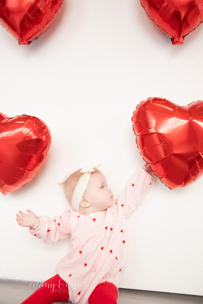 baby girl sits in front of white wall decorated with red heart balloons for valentine's day and reaches up for balloons