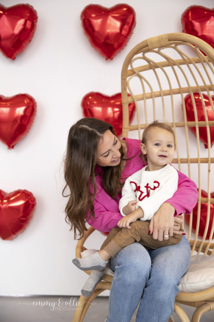 toddler boy sits in a decorative chair with his mama in front of wall decorated with red heart balloons for valentines day