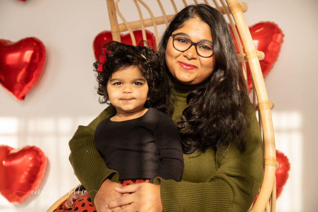 toddler girl sits on mother's lap in decorative chair in front of wall decorated with red heart balloons for valentine's day