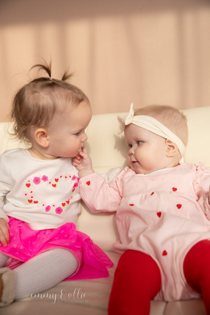 baby girls sit on couch while one reaches up and touches the other's lips