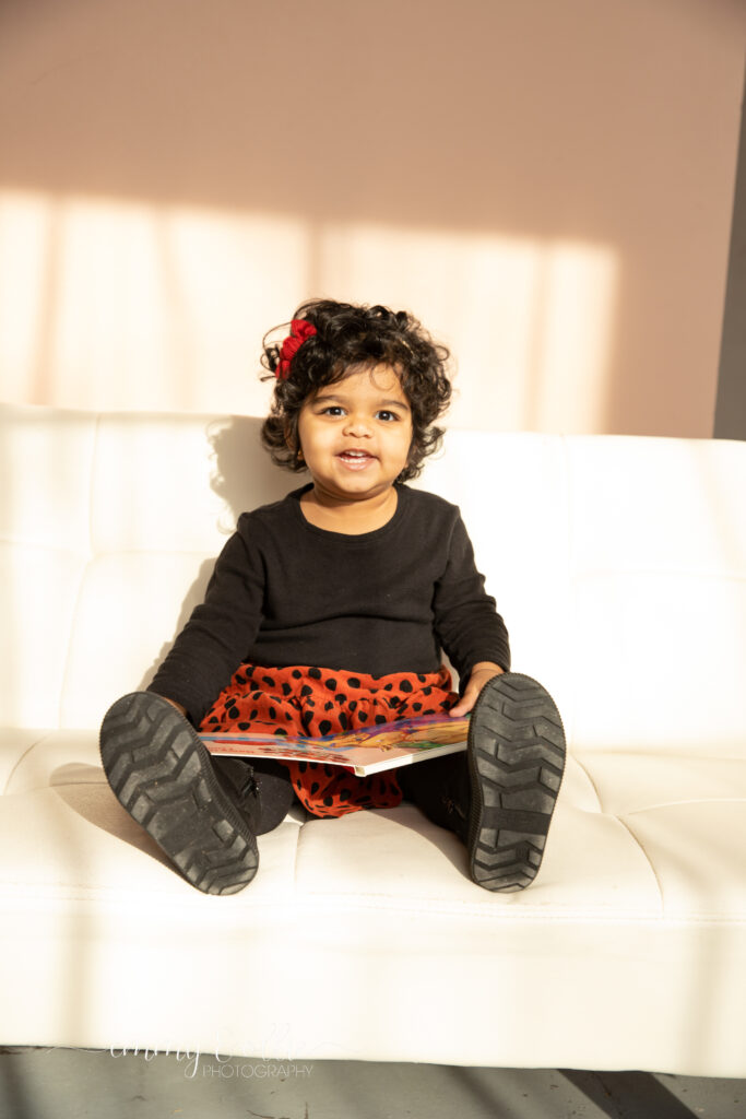 toddler girl sits and reads a book on white couch in front of pink wall