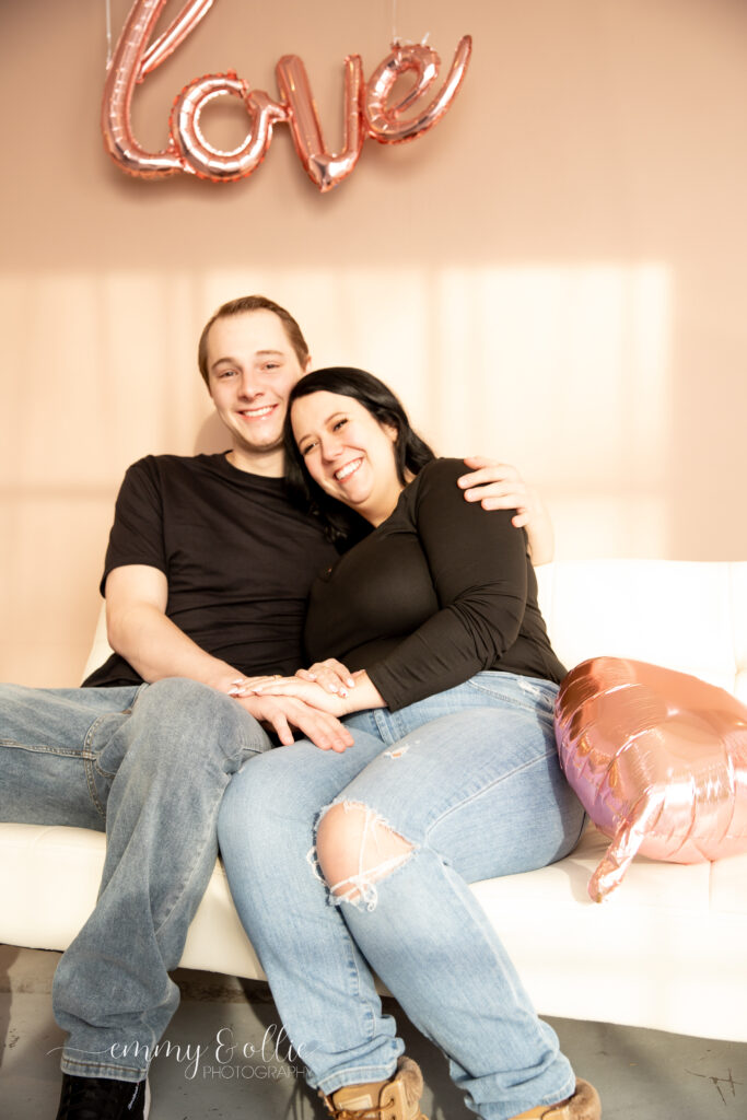 woman and man snuggle together on white couch with a pink heart balloon in front of pink wall decorated with pink love balloon