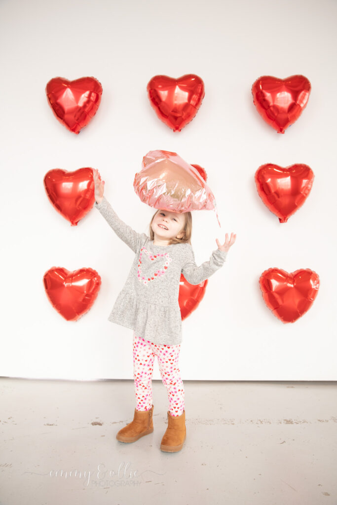 toddler girl tosses up pink heart balloon in front of white wall decorated with red heart balloons for valentine's day