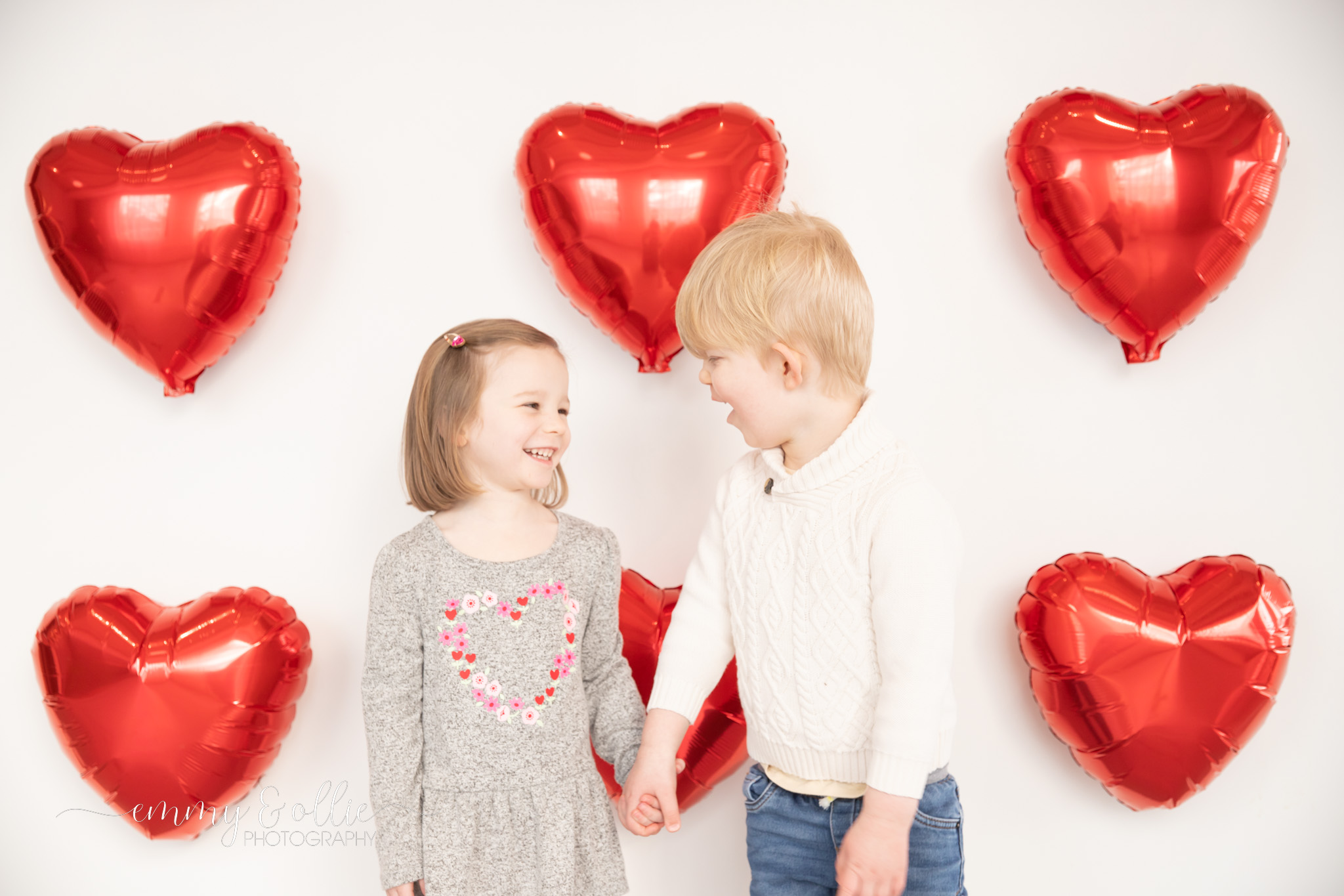 toddler girl and boy hold hands and laugh in front of white wall decorated with red heart balloons for valentine's day