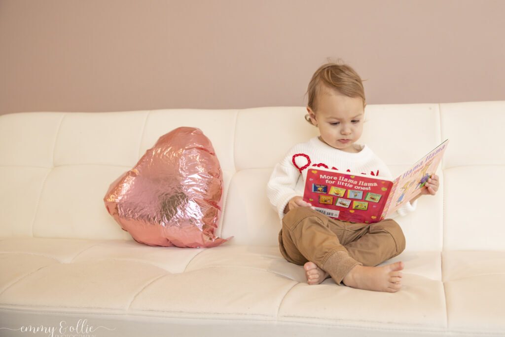 Toddler boy sits on a white couch in front of a pink wall with a pink heart balloon next to him while he reads llama llama I love you with a quizzical look on his face