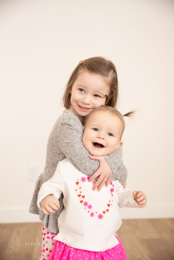 toddler sister hugs baby sister around neck as they both smile