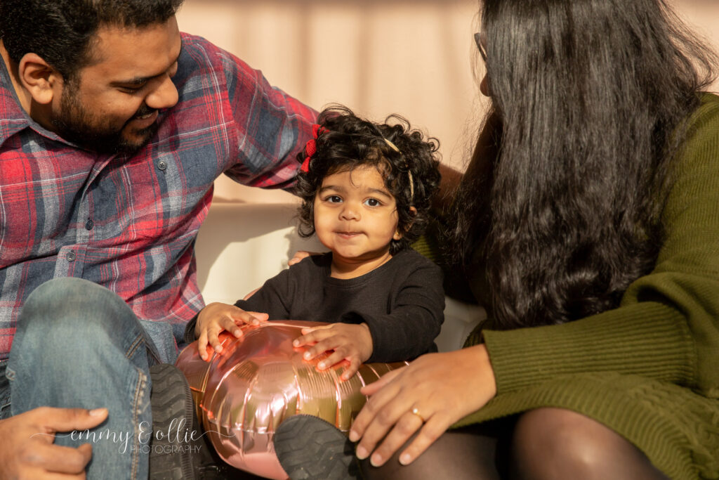 toddler girl holds pink heart balloon as parents look at her and smile