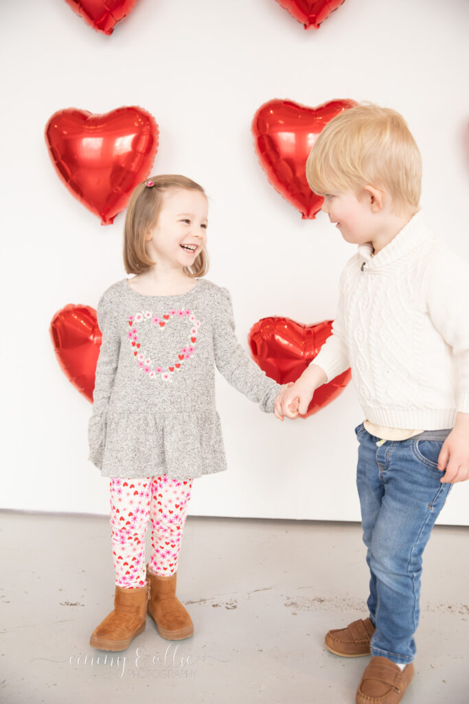 toddler girl and boy hold hands and laugh in front of white wall decorated with red heart balloons for valentine's day