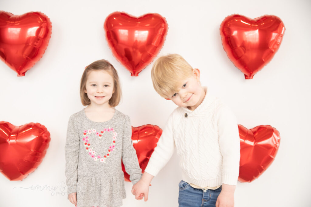 toddler girl and boy hold hands in front of white wall decorated with red heart balloons for valentine's day