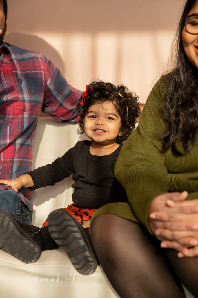 toddler girl sits on couch between parents and smiles