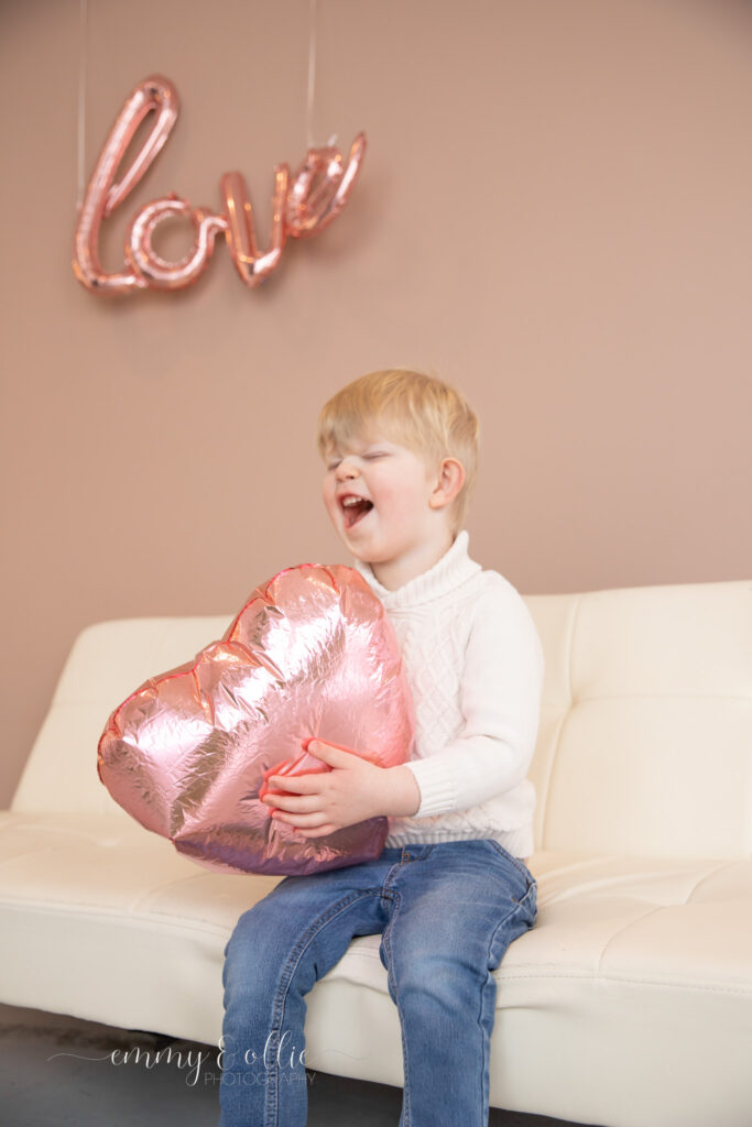 toddler boy laughs while sitting on white couch holding pink heart balloon in front of pink wall with pink love balloon