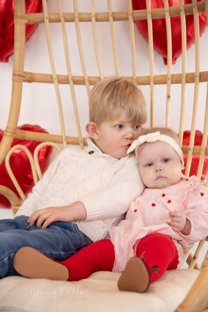 toddler boy and baby sister sit on decorative wooden chair while brother kisses sister on head in front of white wall decorated with red heart balloons for valentine's day