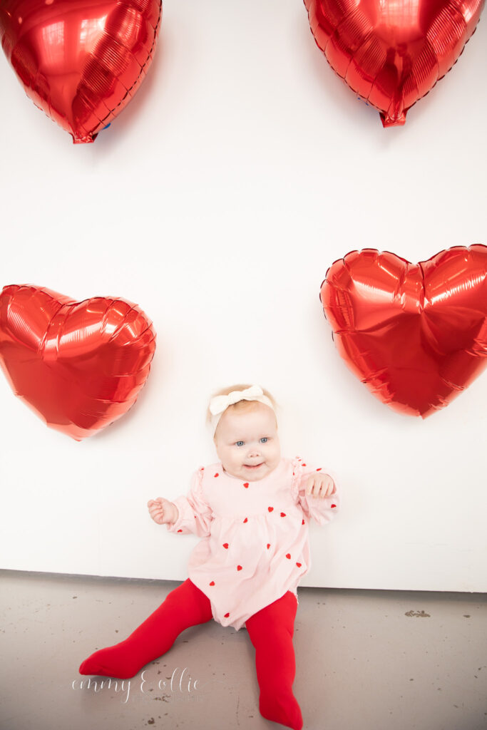 baby girl sits in front of white wall decorated with red heart balloons for valentine's day