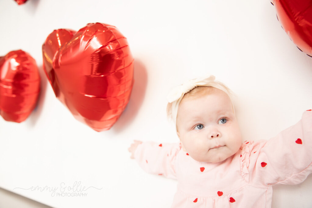 baby girl sits in front of white wall decorated with red heart balloons for valentine's day and reaches for the balloons