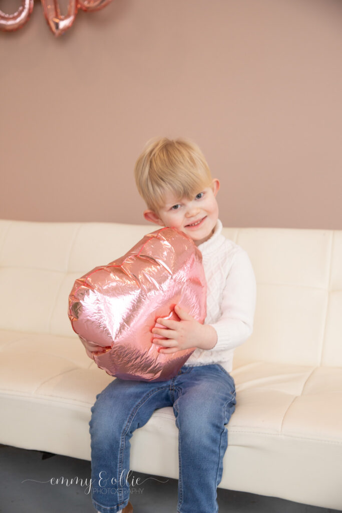 toddler boy sits on white couch holding pink heart balloon and smiles at camera