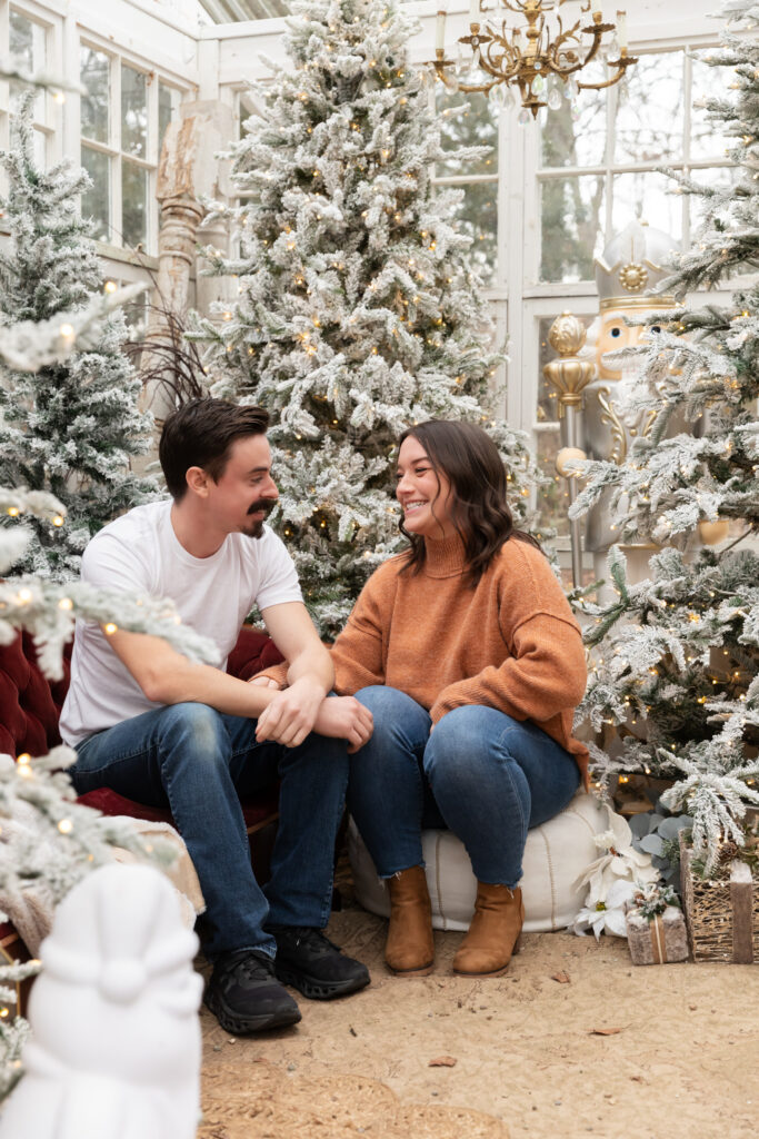 couple laughs and looks at each other surrounded by snow flecked christmas trees in the greenhouse at Rustic Wild Arrow in brooklyn michigan