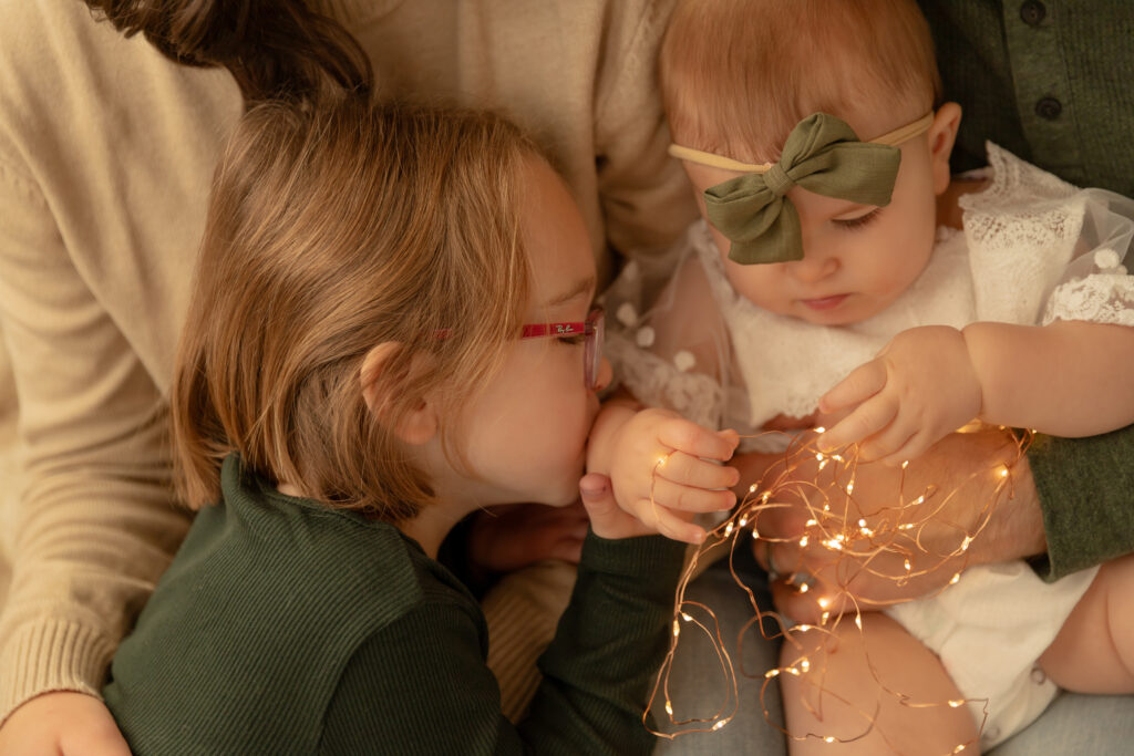 young girl kisses baby sister's hand while cuddling with parents and baby holds copper wired string lights at studiostudio in ann arbor michigan