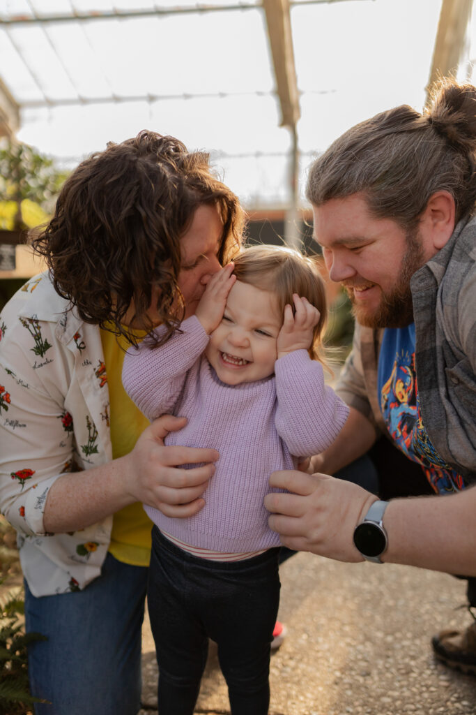 toddler girl smiles and laughs as parents lean in for kisses on sides of her head in the conservatory at matthaei botanical gardens in ann arbor michigan