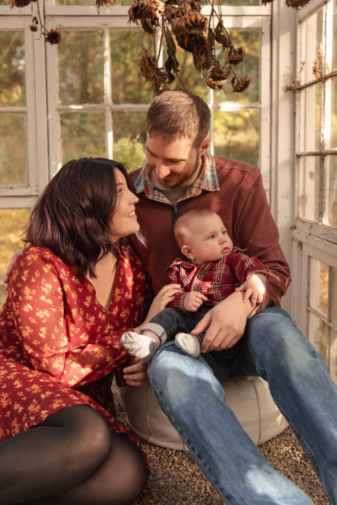 family of three sits on poof in greenhouse, mother looks up to father, baby looks outside at rustic wild arrow in brooklyn michigan