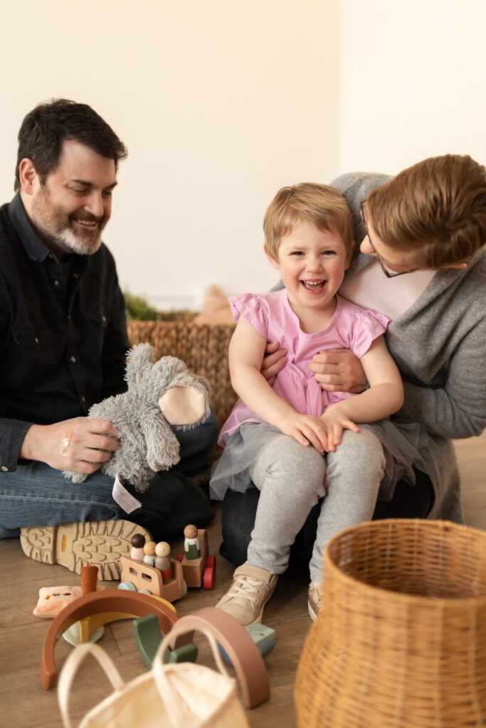 young girl sits on lap of mother laughing and playing while her father holds her elephant toy in studiostudio in ann arbor michigan