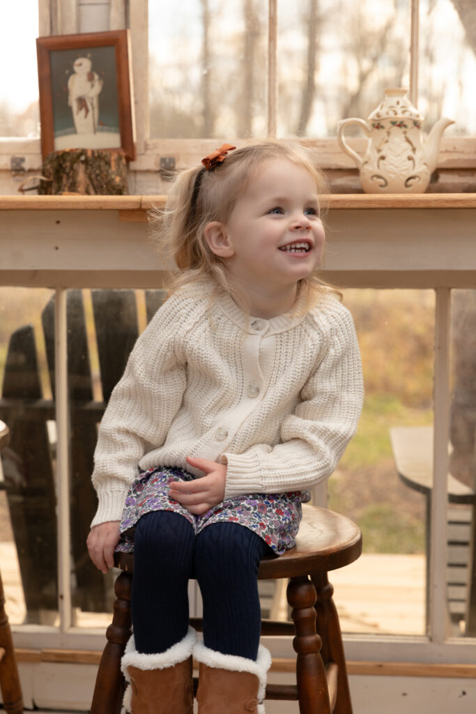 young girl sits on stool and smiles off camera in greenhouse decorated for christmas at fern and pine in dexter michigan