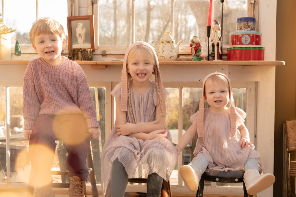 three young siblings smile at camera with twinkle lights in foreground in greenhouse decorated for christmas at fern and pine in dexter michigan