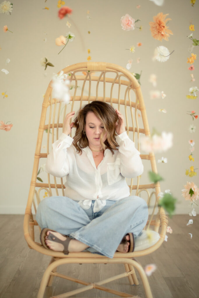 woman sits in rattan chair with floating flowers all around, adjusting hair and looking down to ground at studiostudio in ann arbor michigan