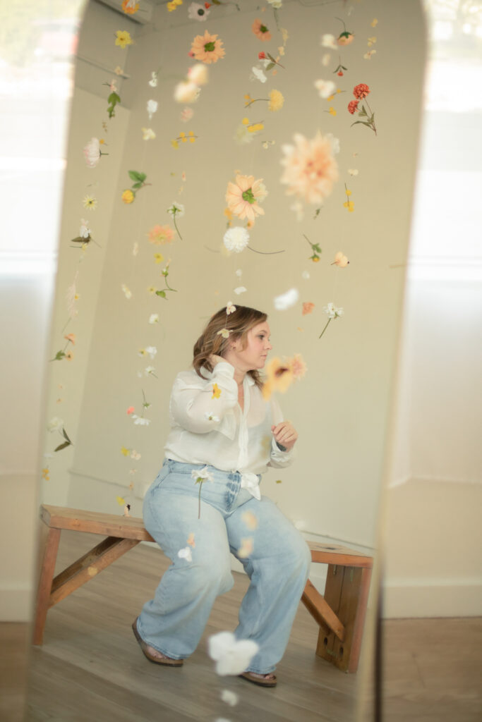 mirror reflection of woman sitting on bench with floating flowers all around, adjusting hair at studiostudio in ann arbor michigan