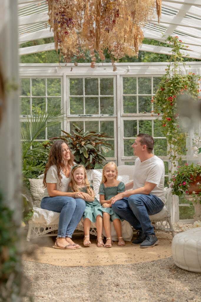 family of four sits on white wicker sofa in greenery filled greenhouse at rustic wild arrow in brooklyn michigan