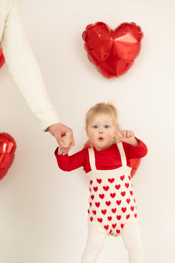 toddler girl holds hands with father and points towards camera in front of white wall decorated with red heart balloons at studiostudio in ann arbor michigan