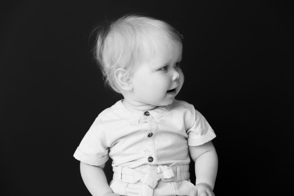 Portrait of a baby girl in black and white against a black background as she looks off camera to the right of frame