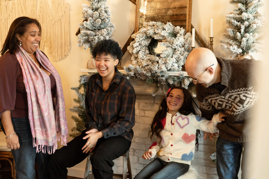 Family of 4 laugh together in front of fireplace decorated for christmas