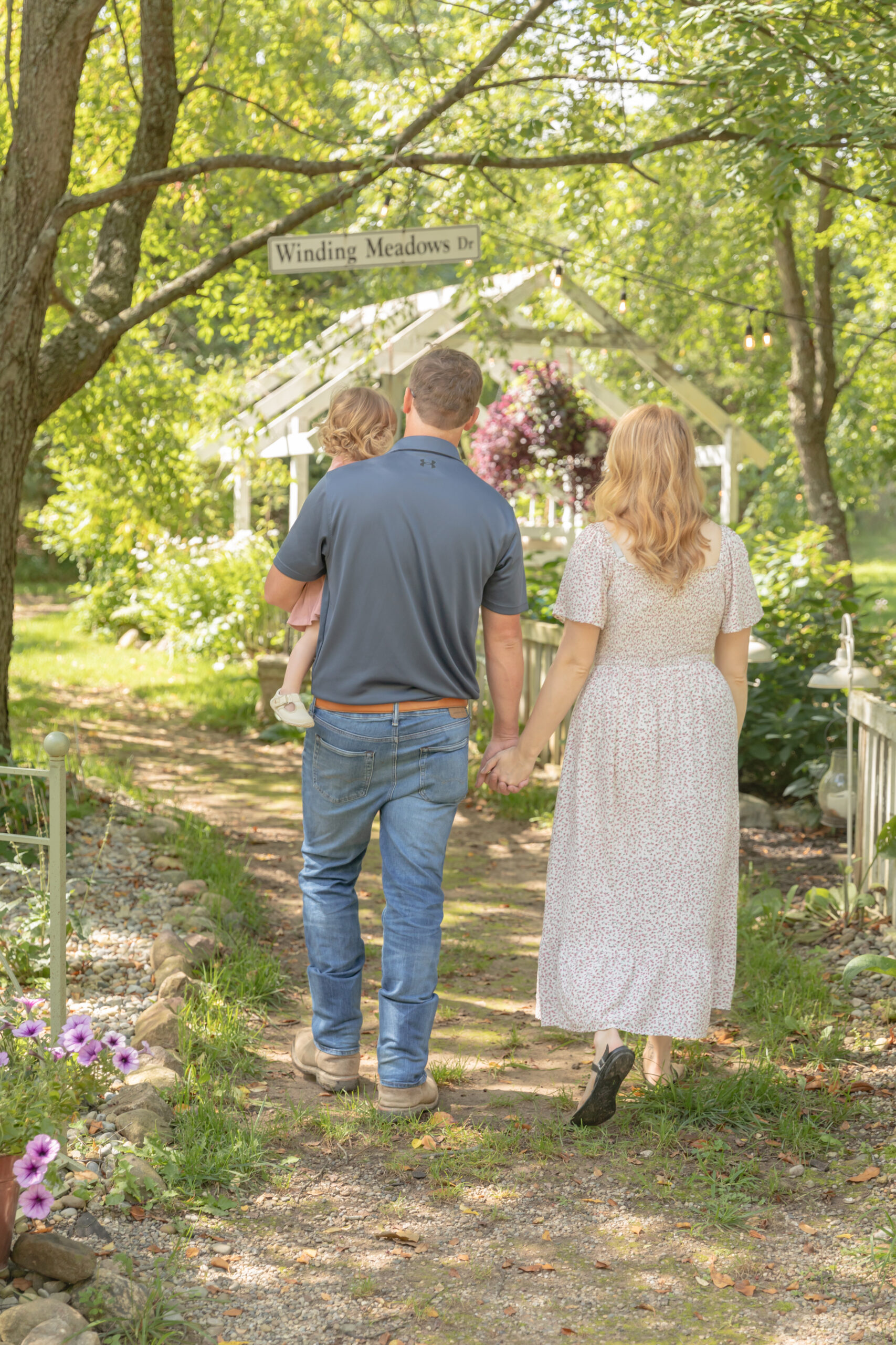 Mother and father walk hand in hand while carrying toddler girl along a dirt path in a garden paradise at Rustic Wild Arrow in Brooklyn, Michigan