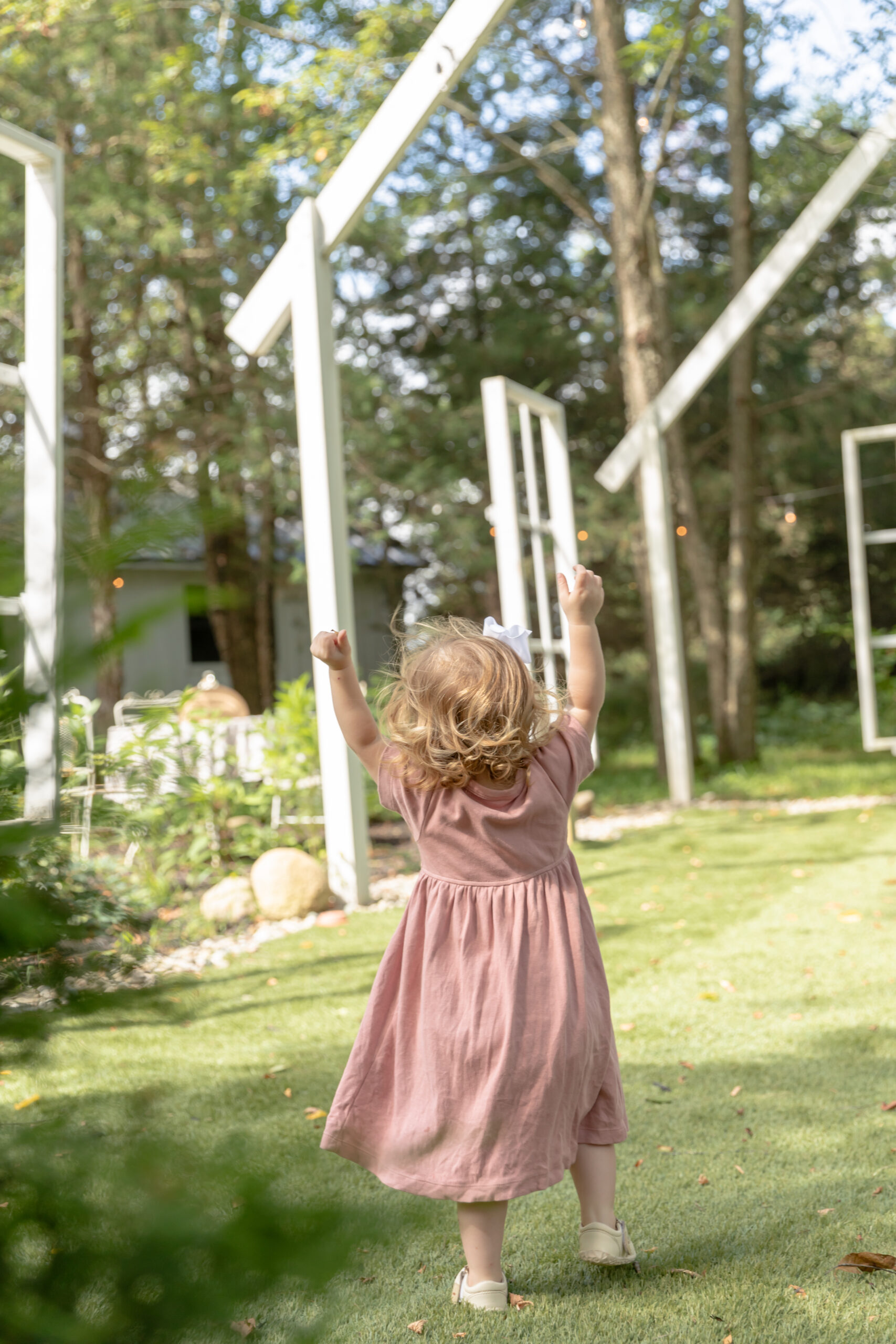 Toddler girl throws her hands in the air as she walks away from the camera in an open air chapel at Rustic Wild Arrow in Brooklyn, Michigan