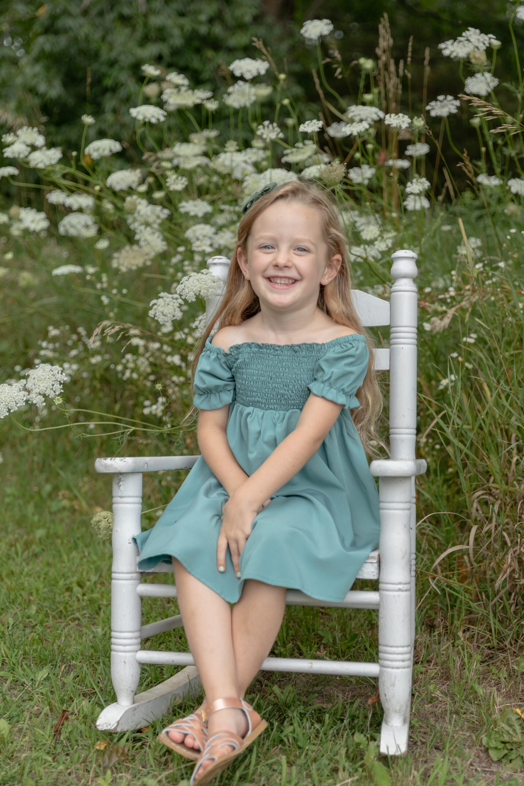 Young girl poses in a small white rocking chair in a field of Queen Anne's Lace at Rustic Wild Arrow in Brooklyn, Michigan