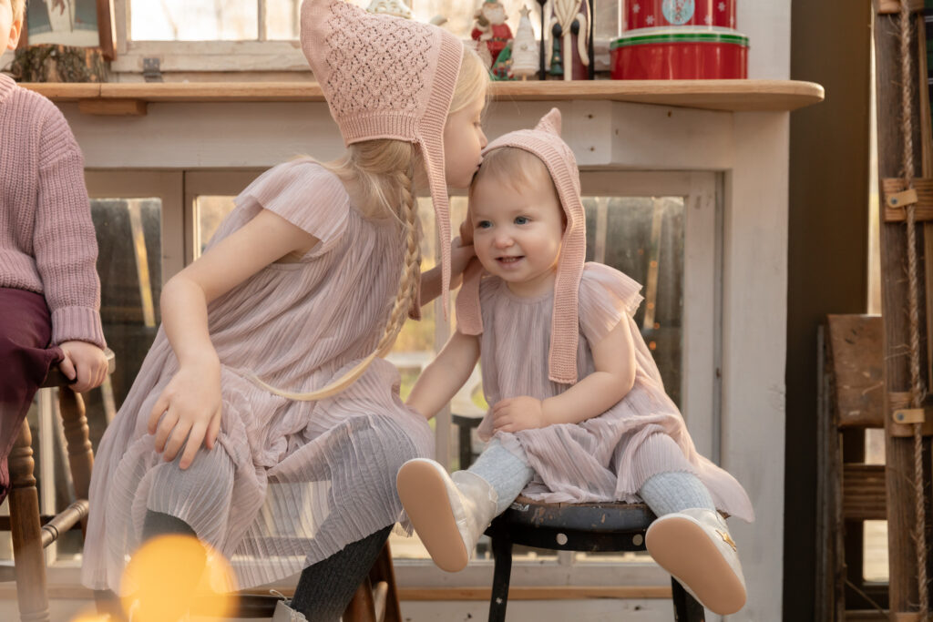 Older sister leans over to give baby sister a kiss on the head while sitting on stools in a greenhouse decorated for Christmas